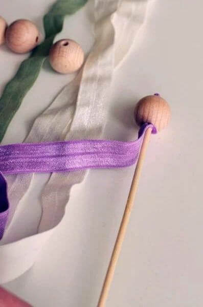 Cắt sợi chun thành một đoạn dài khoảng 16cm, ghim que xiên gỗ vào một đầu dây và luồn qua chiếc lỗ nhỏ của hạt gỗ (Nguồn: Internet)